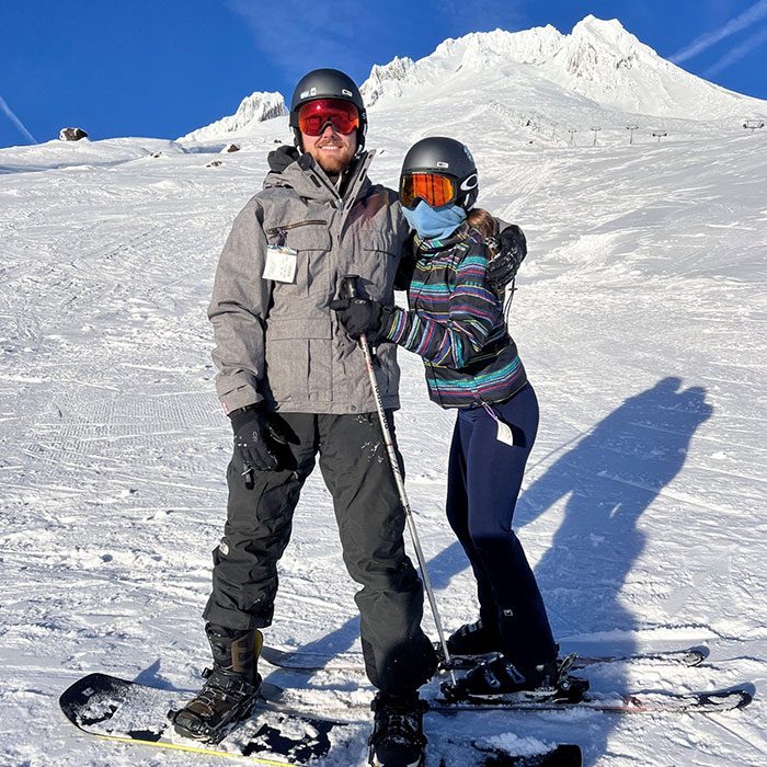 Elizabeth Soldwedel skiing with a companion on a snowy mountain slope, both wearing helmets and ski gear, smiling in the sunshine.