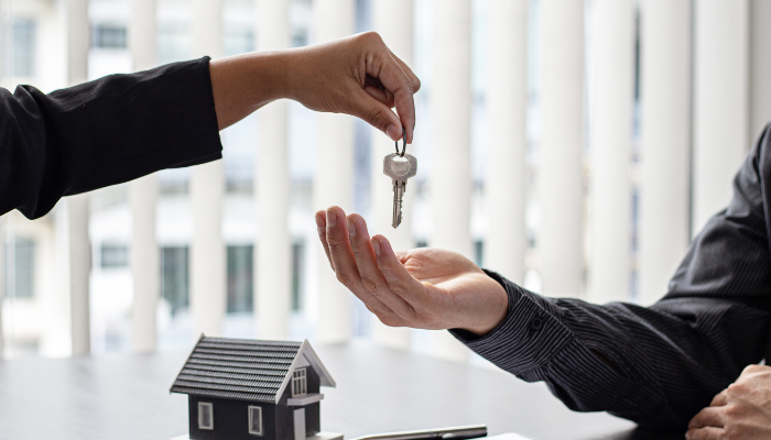 Close-up of a person handing over a house key to another person, with a small model house on a desk in the foreground.