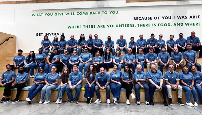 A large group of Draper and Kramer employees in matching blue t-shirts sit together on tiered benches inside a bright space with motivational words on the wall, posing for a team photo during a volunteer or company event.