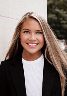 Professional headshot of Elizabeth Soldwedel smiling, wearing a black blazer and white top, standing outdoors against a light-colored wall.