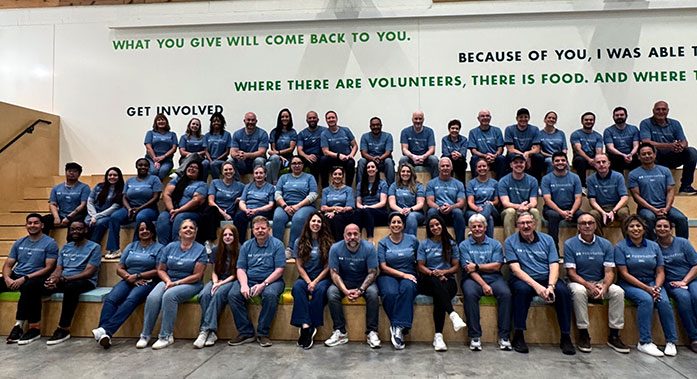Large group of Draper and Kramer employees wearing matching blue volunteer shirts, seated on tiered steps at a community food bank with motivational quotes on the wall behind them.