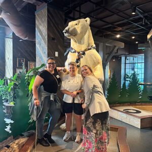 Three women smiling and posing with mini golf putters in front of a large polar bear statue inside an indoor mini golf course.
