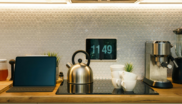 A modern kitchen setup featuring a sleek espresso machine, a stainless-steel kettle, stacked white mugs, a tablet displaying the time, and a laptop — illustrating a connected smart home environment.