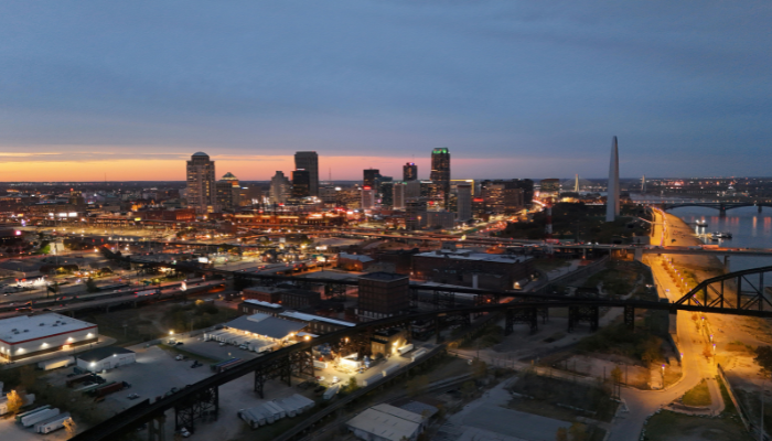 Aerial view of downtown St. Louis, Missouri, at sunset, featuring the illuminated city skyline, the Gateway Arch, the Mississippi Riverfront, and surrounding bridges.