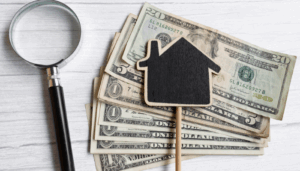 Magnifying glass and small house icon resting on a stack of U.S. dollar bills, symbolizing real estate income and rental costs.