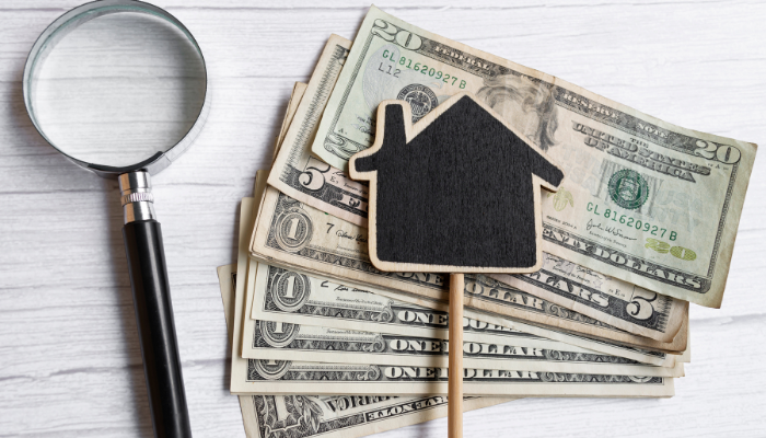 Magnifying glass and small house icon resting on a stack of U.S. dollar bills, symbolizing real estate income and rental costs.