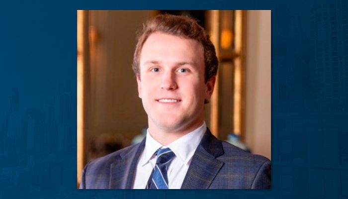 A young adult man with short light-brown hair, wearing a blue suit jacket, white shirt, and striped tie, poses indoors in front of softly lit, elegant background decor.