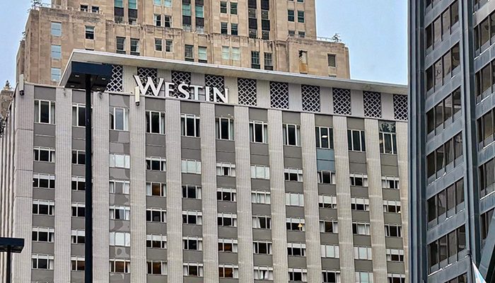 Street-level view of the Westin Michigan Avenue hotel in Chicago, showing the multi-story facade with the Westin signage, surrounding high-rise buildings, leafless trees decorated with lights, and U.S. and Chicago flags in the foreground.