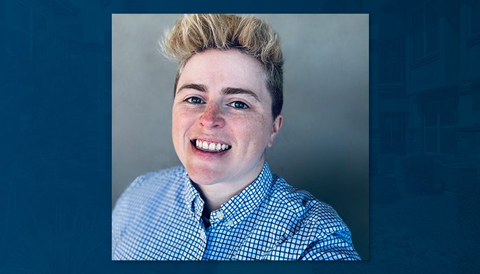 Portrait of a smiling person with short, blond hair wearing a blue checkered button-down shirt, posed against a neutral background.
