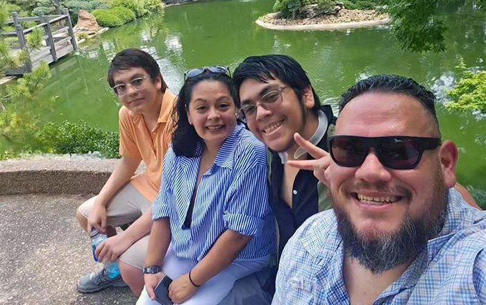 Ida Valdez sitting with her family near a pond; the group smiles at the camera while seated on a walkway with green water and trees in the background.