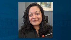 Headshot of Ida Valdez, a woman with shoulder-length dark hair, smiling slightly while seated in an office. She is wearing a black Draper and Kramer shirt, and a framed artwork is visible in the background.