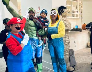 Group of people in costumes at a property event; Ida Valdez is dressed as Mario and holding a microphone, standing next to others dressed as the Hulk, Captain America, a Minion, and a mummy in a lobby area.
