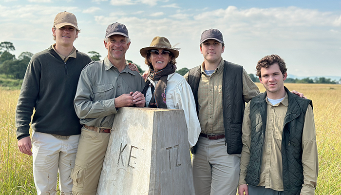 Five people stand together outdoors in a grassy field beside a stone marker, dressed in casual outdoor clothing and hats.