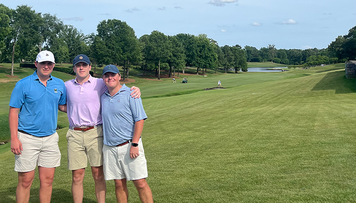 Three men stand together on a golf course with arms around each other, with a wide fairway, trees, and a water feature in the background.