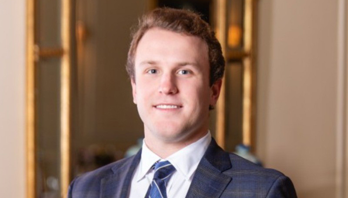 Headshot of a young professional man in a suit and tie, smiling, with a softly blurred indoor office background.