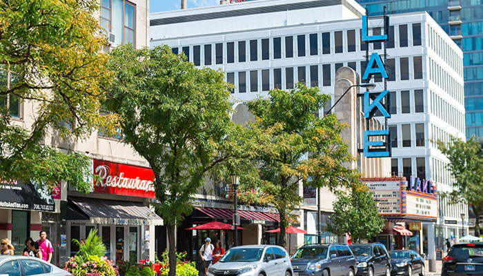 A busy street in downtown Oak Park with shops, restaurants, parked cars, and the Lake Theatre marquee visible among trees and mid-rise buildings.