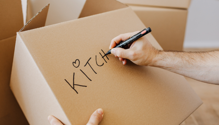Close-up of a person writing “Kitchen” with a marker on a cardboard moving box, with other boxes in the background.