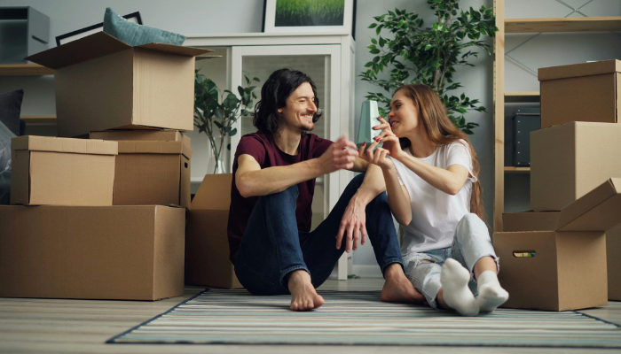 A young couple sits on the floor of their new home surrounded by moving boxes, smiling and talking as they take a break from unpacking.
