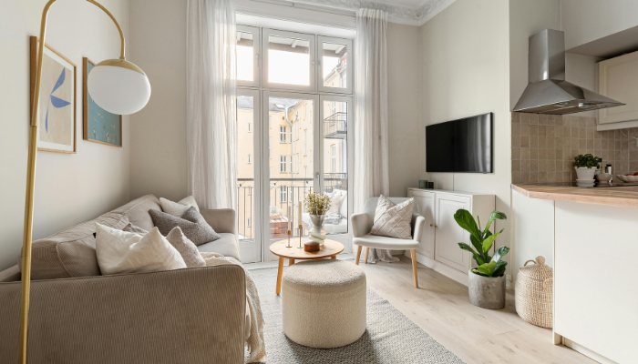 Bright, furnished apartment living area with a neutral-toned sofa, accent chair, round ottoman, and small coffee table positioned near tall windows. The space features light wood flooring, soft natural light, a wall-mounted television, indoor plants, and an adjacent modern kitchen with white cabinetry and stainless steel appliances.