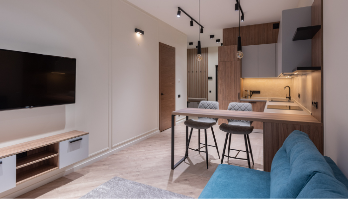 Modern open-concept apartment interior featuring a compact kitchen with wood cabinetry, a breakfast bar with two stools, a blue sofa, wall-mounted television, light wood flooring, and contemporary pendant and track lighting.