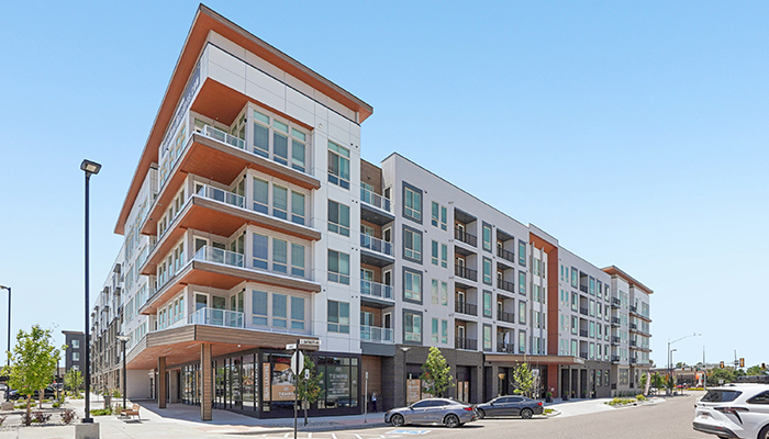 Street-level exterior view of Tempo Nine Mile Station, a modern five-story apartment community in Aurora, Colorado, featuring white and gray façades, glass balconies, ground-floor retail space, and landscaped sidewalks at the corner of a city intersection.