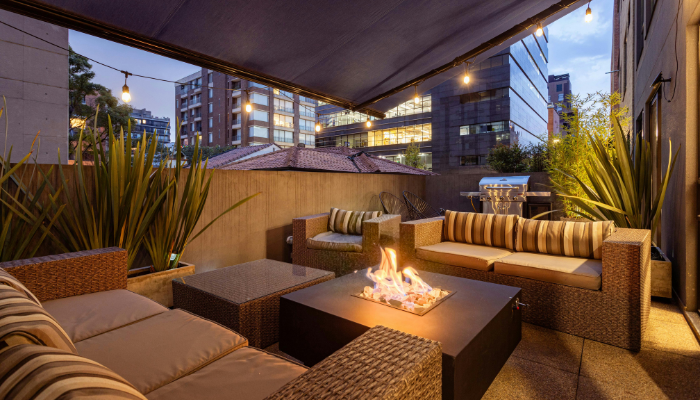 A furnished apartment patio at dusk features woven outdoor seating surrounding a modern gas fire table, with string lights overhead, tall planters, and a city skyline in the background.
