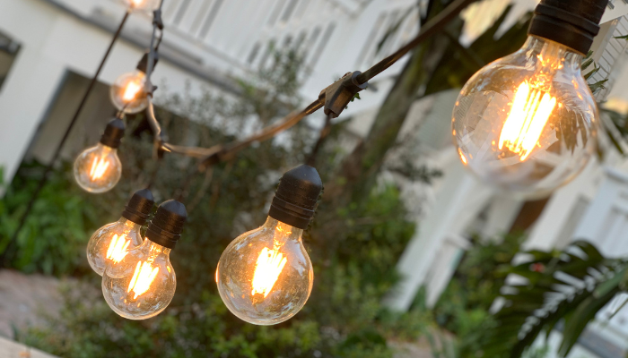 Warm Edison-style string lights hang above a patio, casting a soft golden glow against a backdrop of lush greenery and a residential building, creating a cozy outdoor atmosphere for summer evenings.