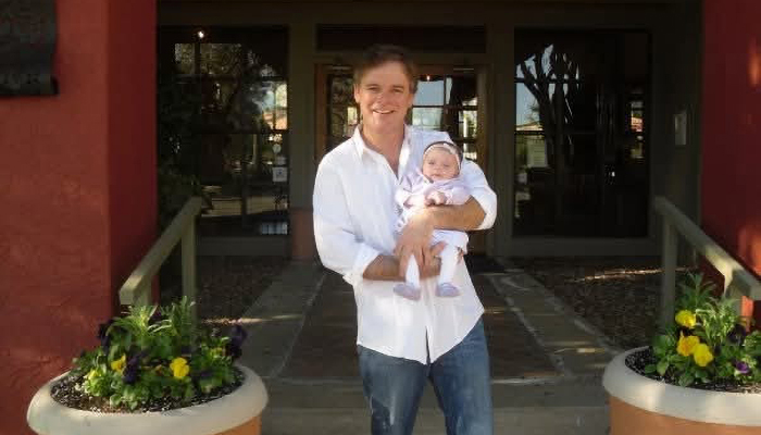 Dallas Comstock smiling while holding his infant child outside a residential building.