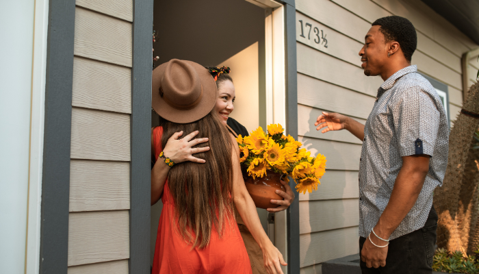 Two friends greet a woman holding a bouquet of sunflowers at the front door of a new home.