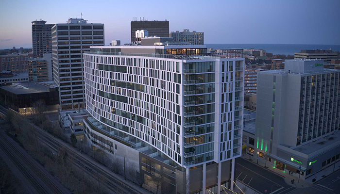 Dusk exterior view of Eva, a 15-story, 268-unit luxury apartment community in downtown Evanston, Illinois, featuring modern glass architecture, illuminated rooftop amenities and Lake Michigan in the background.