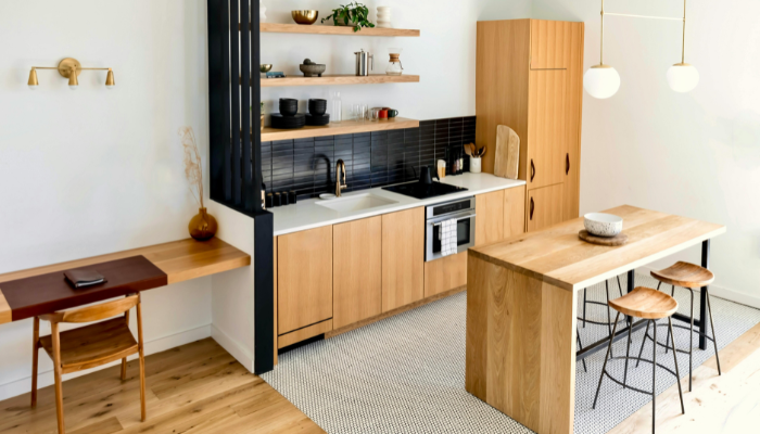 Compact apartment kitchen with light wood cabinetry, open shelving, a narrow island with stools, and a small built-in desk.