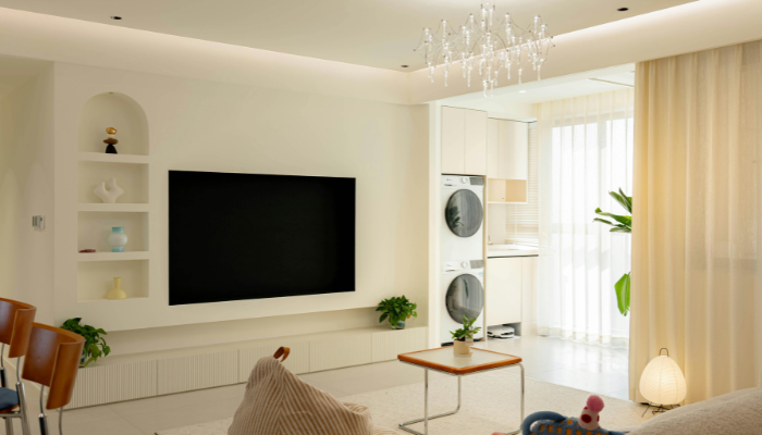Bright apartment living room with light-colored furnishings, a wall-mounted TV, built-in shelving, large windows and an open kitchen with stacked washer and dryer.